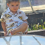 Tudor is registered to the contest to win money with this photo: child, boy, outdoor, sitting, shirt, lion_king, shorts, reflection, glass_table, daylight, plants, stone, curious, casual_clothing, young, portrait, sunlight, shadow, concrete, relaxed