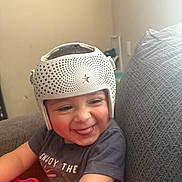 Ezekiel is registered to the contest to win money with this photo: baby, child, infant, helmet, protective_helmet, smile, tongue_out, toy_piano, xylophone, tshirt, arm, couch, pillow, living_room, indoor, happy, playful, cheerful, decor, portrait