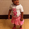 toddler, girl, child, smiling, standing, pink_skirt, white_tshirt, pigtails, beads, indoor, tile_floor, brick_wall, shoes, happy, cute, fashion, portrait, person, young_child, casual_clothing