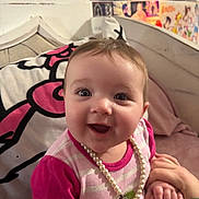 Violet joined the competition — help win amazing prizes! baby, child, smiling, pearl_necklace, pink_clothing, tattooed_hands, bed, pillow, colorful_background, portrait, happy, cute, infant, person, indoor, holding_hands, face, young_child, joyful, cozy