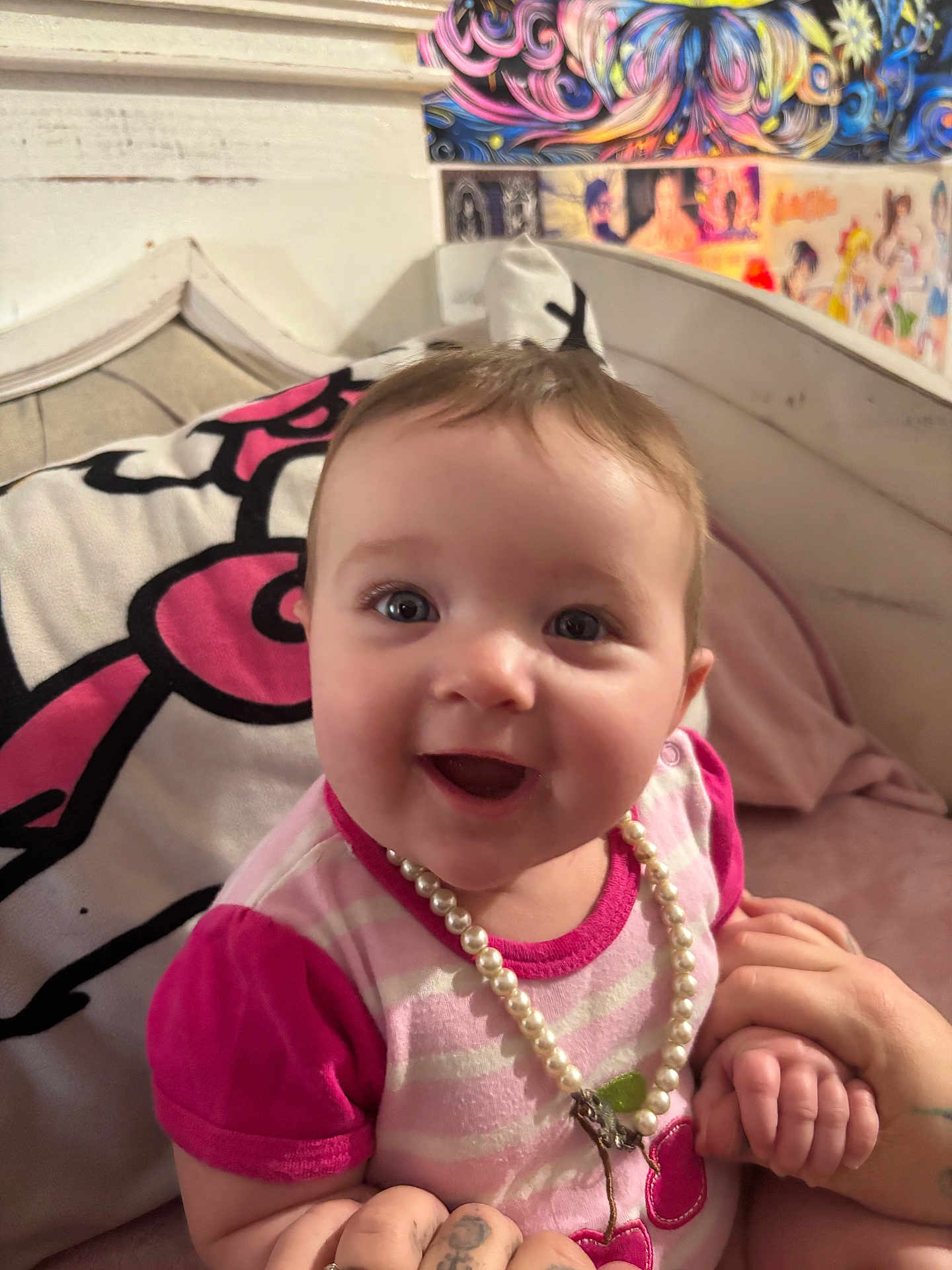 Violet joined the competition — help win amazing prizes! baby, child, smiling, pearl_necklace, pink_clothing, tattooed_hands, bed, pillow, colorful_background, portrait, happy, cute, infant, person, indoor, holding_hands, face, young_child, joyful, cozy