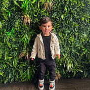 Dayron is registered to the contest to win money with this photo: toddler, child, boy, standing, greenery, plants, leafy_background, jacket, black_pants, sneakers, indoor, portrait, smiling, cute, fashion, casual_clothing, floor, wooden_floor, happy, person