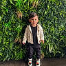 toddler, child, boy, standing, greenery, plants, leafy_background, jacket, black_pants, sneakers, indoor, portrait, smiling, cute, fashion, casual_clothing, floor, wooden_floor, happy, person