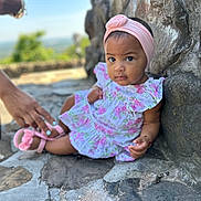 Ariella joined the competition — help win amazing prizes! baby, child, girl, headband, pink, dress, outdoor, stone_wall, sandal, hand, helping_hand, curious, expression, sitting, portrait, footwear, nature, blurred_background, person, cute