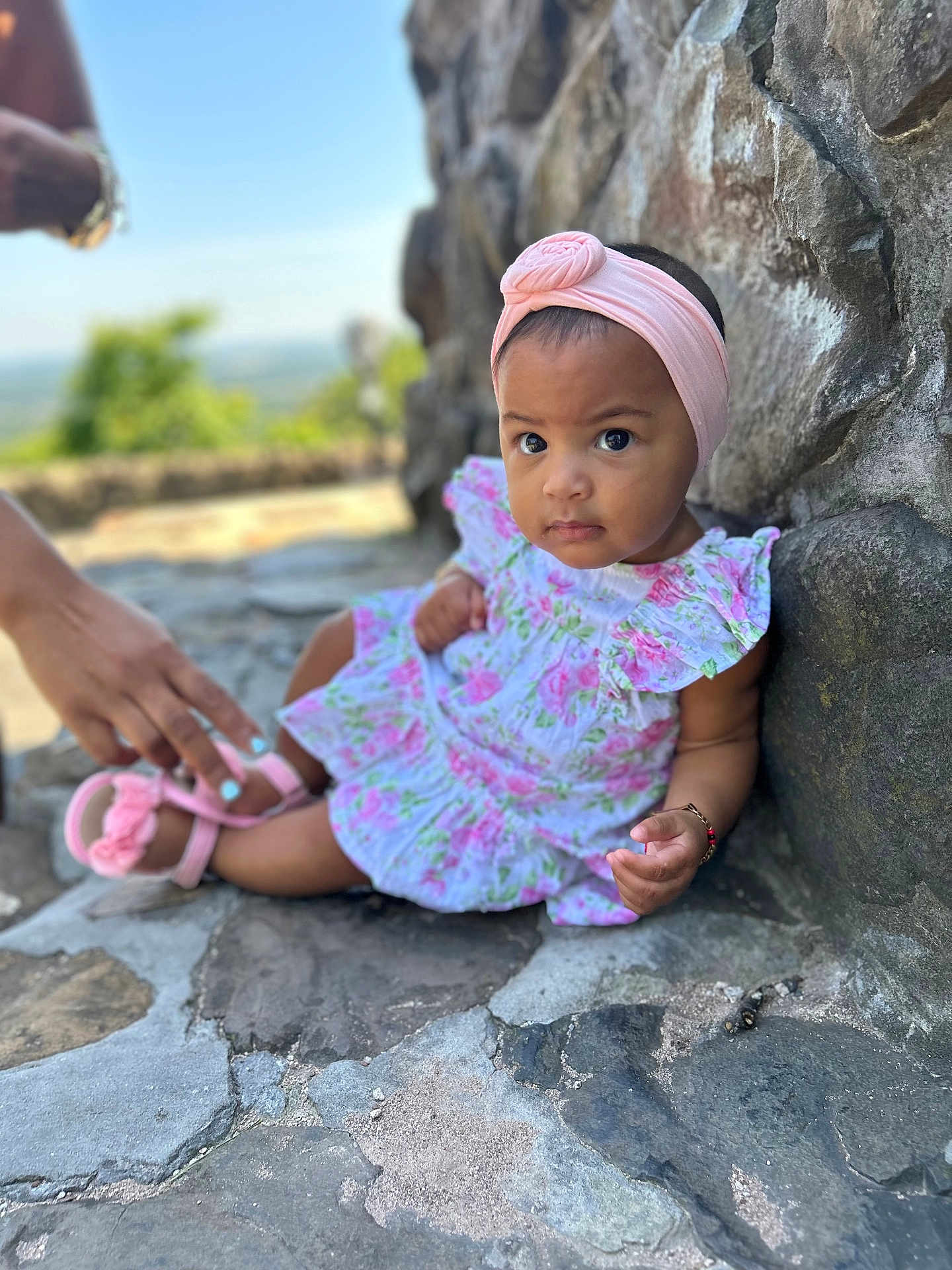 Ariella joined the competition — help win amazing prizes! baby, child, girl, headband, pink, dress, outdoor, stone_wall, sandal, hand, helping_hand, curious, expression, sitting, portrait, footwear, nature, blurred_background, person, cute