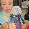 toddler, child, blue_eyes, bib, smoothie, sippy_cup, carpet, toy, indoor, baby, clothing, hand, face, sitting, rosy_cheeks, hair, drink, plastic_cup, household, comfort