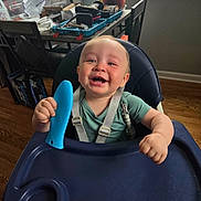 Christopher is registered to the contest to win money with this photo: baby, high_chair, smiling, toy, indoor, child, happy, seat, face, furniture, plant, window, wood_floor, green_shirt, straps, home, daylight, expression, holding, infant