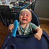 baby, high_chair, smiling, toy, indoor, child, happy, seat, face, furniture, plant, window, wood_floor, green_shirt, straps, home, daylight, expression, holding, infant