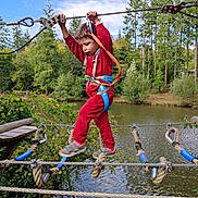 Emilio participe au concours pour gagner de l'argent avec cette photo : child, rope_bridge, adventure, safety_harness, outdoor, forest, pond, trees, sky, clothing, red_outfit, rope, wooden_platform, nature, park, active, young_child, play, balance, daytime