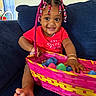 Athena joined the competition — help win amazing prizes! toddler, child, smiling, basket, plastic_eggs, beaded_hair, braids, red_shirt, couch, indoor, happy, barefoot, bracelets, colorful, toy, person, portrait, home, cute, playful