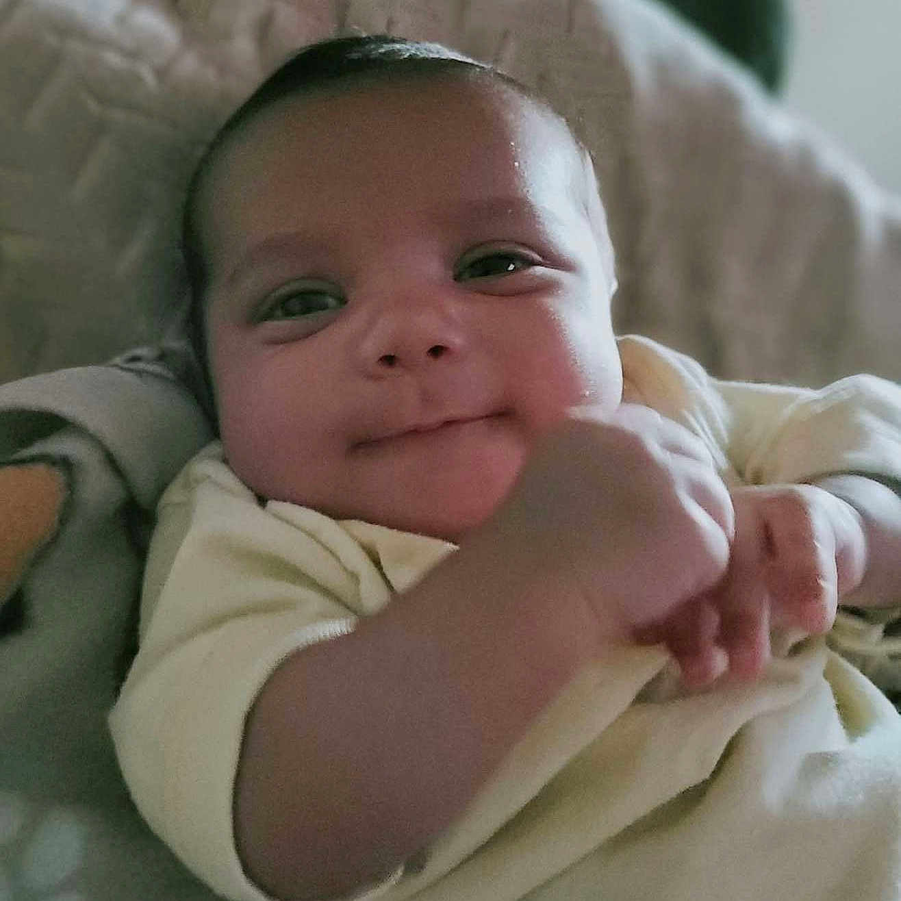 Chief is registered to the contest to win money with this photo: baby, infant, child, smiling, portrait, close_up, lying_down, onesie, clothing, arm, face, eyes, mouth, head, blanket, cushion, bed, indoor, cozy, photography