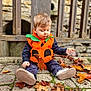 autumn_leaves, baby_shoes, casual_clothing, child, costume, curious, cute, fall, leaf, nature, outdoor, playful, pumpkin_costume, seasonal, sitting, stone_pavement, stone_wall, toddler, wooden_fence, young_child