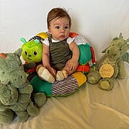 Carlin is registered to the contest to win money with this photo: baby, child, stuffed_animal, cushion, toy, green, dragon, caterpillar, overalls, socks, white_shirt, indoor, seated, expression, cute, young, portrait, soft_toys, colorful, background