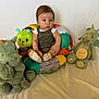 baby, child, stuffed_animal, cushion, toy, green, dragon, caterpillar, overalls, socks, white_shirt, indoor, seated, expression, cute, young, portrait, soft_toys, colorful, background