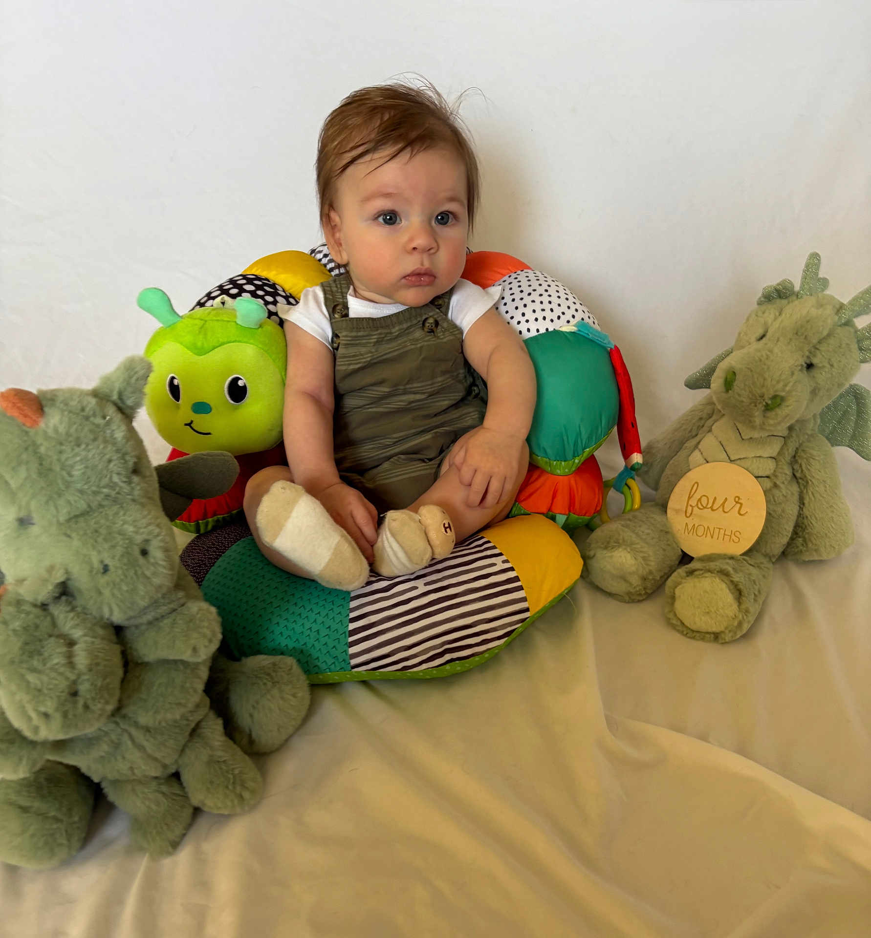 Carlin is registered to the contest to win money with this photo: baby, child, stuffed_animal, cushion, toy, green, dragon, caterpillar, overalls, socks, white_shirt, indoor, seated, expression, cute, young, portrait, soft_toys, colorful, background