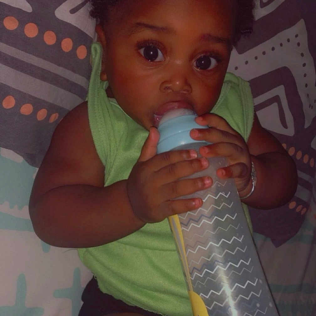 Geneïl a rejoint le concours — aidez-le/la à gagner de superbes lots ! baby, bottle, child, clothing, cute, dark_skin, drinking, expression, face, green_shirt, hand, holding, indoors, infant, lying_down, patterned_blanket, portrait, resting, wide_eyes, young_child