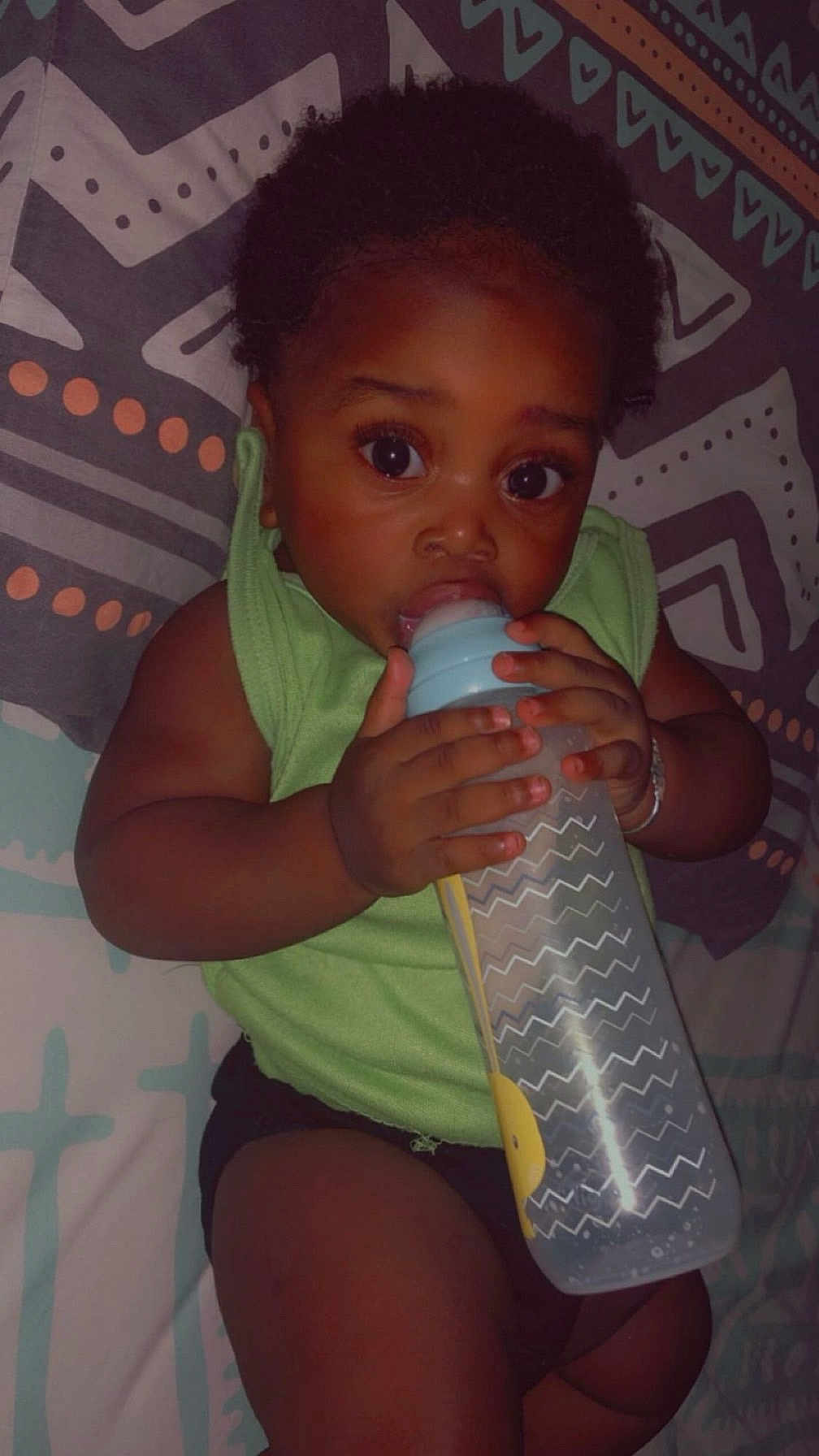 Geneïl a rejoint le concours — aidez-le/la à gagner de superbes lots ! baby, bottle, child, clothing, cute, dark_skin, drinking, expression, face, green_shirt, hand, holding, indoors, infant, lying_down, patterned_blanket, portrait, resting, wide_eyes, young_child