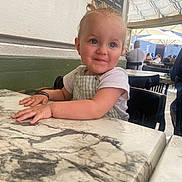 Lya a rejoint le concours — aidez-le/la à gagner de superbes lots ! toddler, child, smile, face, blue_eyes, table, marble, cafe, indoor, person, clothing, short_sleeve, hair, earrings, hand, chair, background, people, daylight, expression