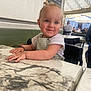 toddler, child, smile, face, blue_eyes, table, marble, cafe, indoor, person, clothing, short_sleeve, hair, earrings, hand, chair, background, people, daylight, expression