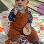 toddler, child, toy, play_mat, overalls, shoes, library, bookshelf, sunlight, indoor, floor, carpet, chewing, clothing, person, sitting, cute, baby, play, colorful