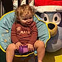 toddler, child, pacifier, purple_cup, curly_hair, blue_chair, yellow_trim, plush_bear, santa_hat, indoor, floor_mat, barefoot, clothing, happy, sitting, toy, holiday, cute, smile, baby