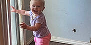 Oaklynn is registered to the contest to win money with this photo: baby, barefoot, child, curtains, exploring, flooring, glass_door, happy, home, indoor, natural_light, person, pink_shorts, purple_shirt, smiling, standing, toddler, vertical_blinds, wooden_floor, young_child