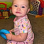 baby, child, onesie, pink_clothing, toy, blue_toy, carpet, indoor, furniture, playroom, cute, face, happy, sitting, childrens_toy, cabinet, floor, smiling, infant, young_child