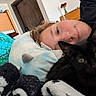 cat, black_cat, person, face, eyes, cuddling, bedroom, bed, pillow, blanket, plush_toy, stuffed_animal, indoor, home, cozy, sleeping, lying_down, portrait, black_fur, green_eyes