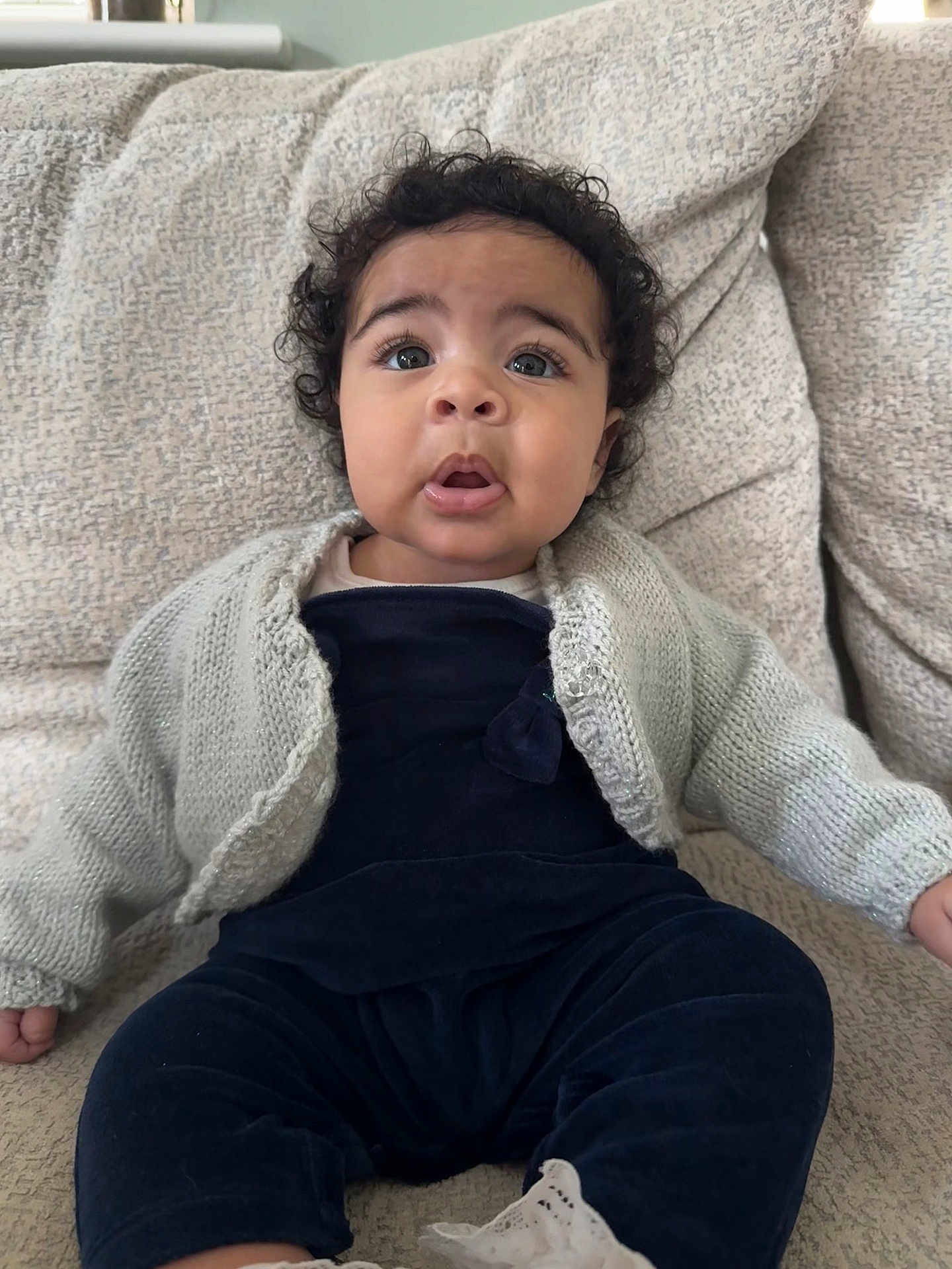Nyah joined the competition — help win amazing prizes! baby, child, curly_hair, blue_eyes, knitted_cardigan, velvet_clothing, couch, cozy, indoor, portrait, sitting, expression, cute, infant, furniture, soft_texture, home, person, young_child, casual_wear