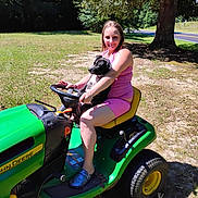 Toni Collins is registered to the contest to win money with this photo: woman, dog, puppy, lawn_tractor, john_deere, outdoor, tree, grass, sunny, summer, casual_clothing, pink_outfit, footwear, pet, animal, nature, rural, smiling, sitting, holding