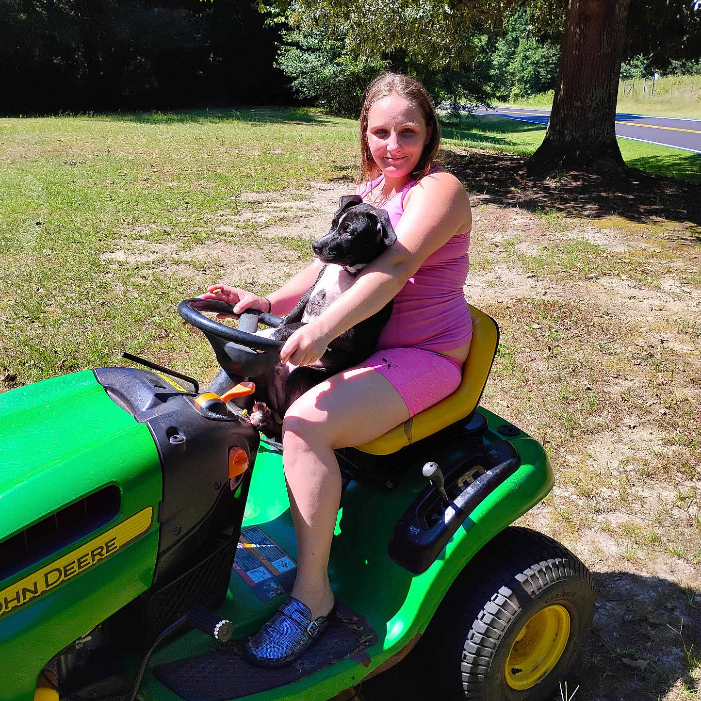 Toni Collins is registered to the contest to win money with this photo: animal, casual_clothing, dog, footwear, grass, holding, john_deere, lawn_tractor, nature, outdoor, pet, pink_outfit, puppy, rural, sitting, smiling, summer, sunny, tree, woman