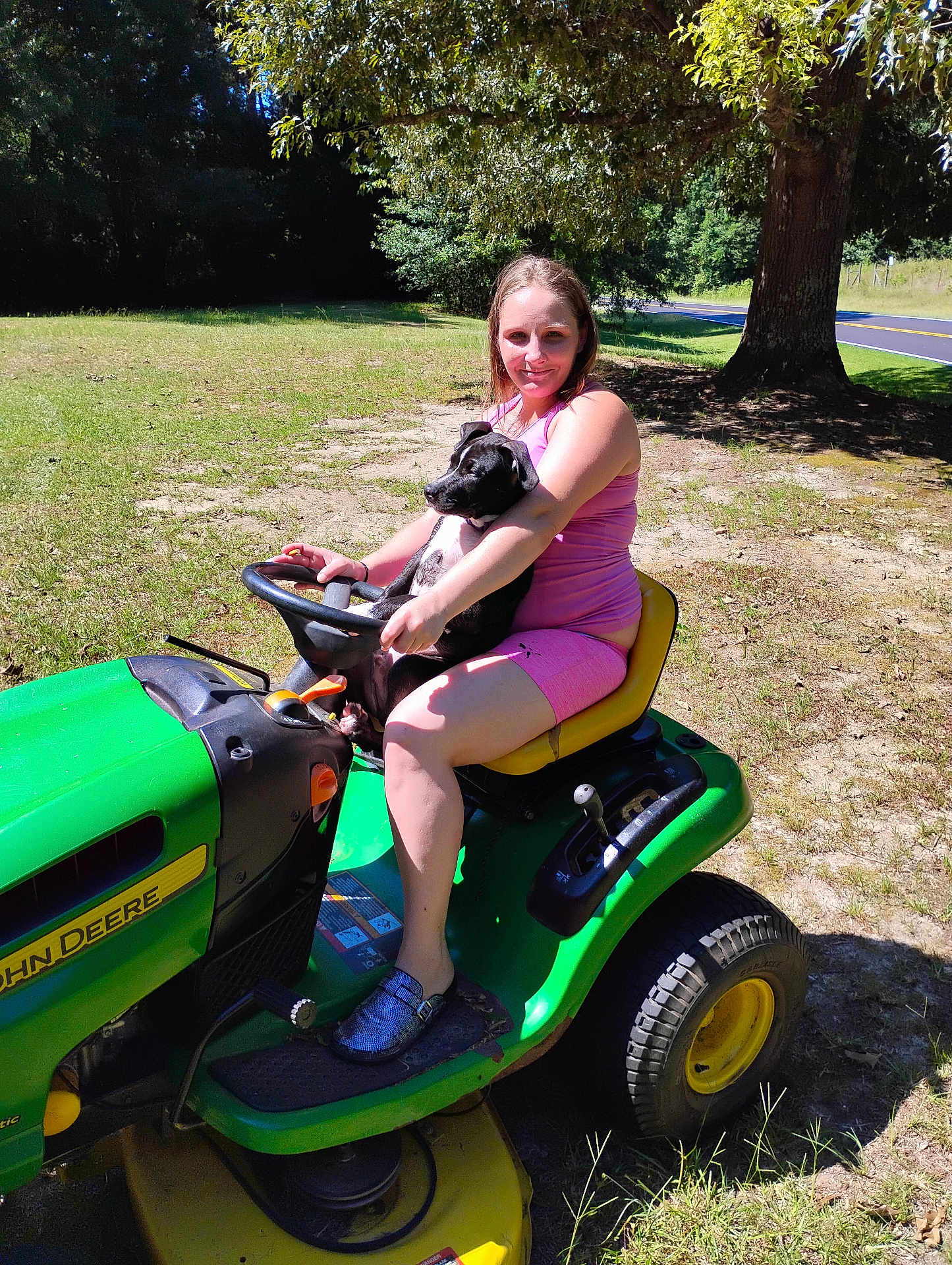 Toni Collins is registered to the contest to win money with this photo: woman, dog, puppy, lawn_tractor, john_deere, outdoor, tree, grass, sunny, summer, casual_clothing, pink_outfit, footwear, pet, animal, nature, rural, smiling, sitting, holding