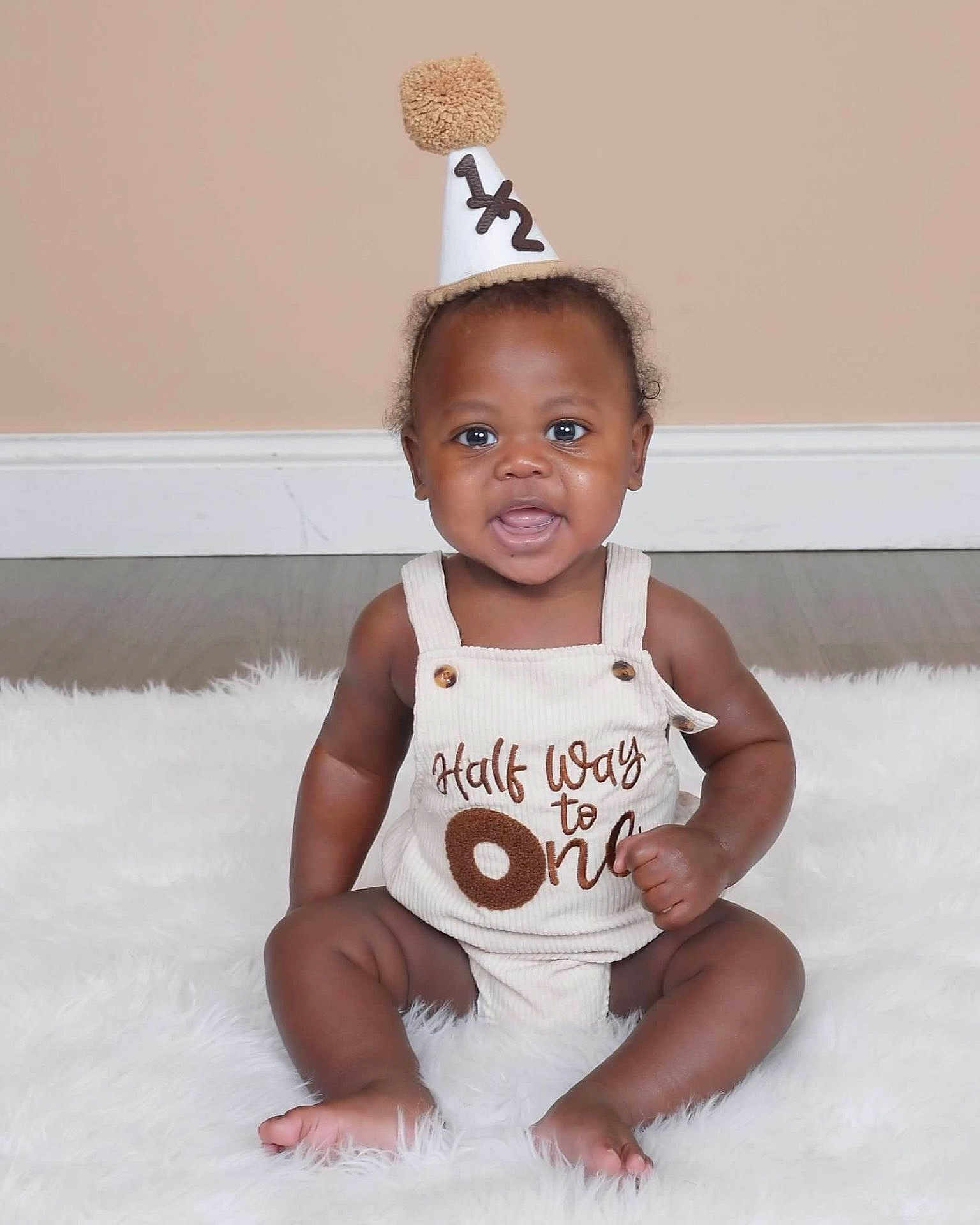Drayson is registered to the contest to win money with this photo: baby, infant, half_birthday, party_hat, romper, sitting, fluffy_rug, smile, cheeks, eyes, portrait, indoor, milestone, celebration, cute, adorable, barefoot, wood_floor, wall_baseboard, one_half