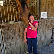 Samira a rejoint le concours — aidez-le/la à gagner de superbes lots ! child, horse, stable, wooden_wall, hay, smiling, pink_shirt, black_shoes, animal, barn, pet, outdoor, friendship, portrait, farm, fence, casual_clothing, young_person, daylight, rustic