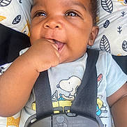 Milo joined the competition — help win amazing prizes! baby, infant, child, car_seat, harness, snoopy_shirt, portrait, face, smiling, hand, fist, chewing, sitting, indoors, patterned_fabric, cushion, black_hair, brown_skin, close_up, safe