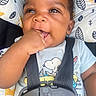 baby, infant, child, car_seat, harness, snoopy_shirt, portrait, face, smiling, hand, fist, chewing, sitting, indoors, patterned_fabric, cushion, black_hair, brown_skin, close_up, safe
