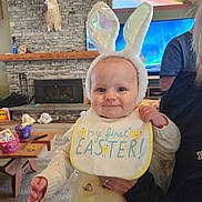 Madelyn is registered to the contest to win money with this photo: baby, bunny_ears, bib, smile, indoor, living_room, fireplace, stone_wall, deer_head, television, person_holding_baby, sweater, dress, carpet, wood_ceiling, table, toys, hand, cute, holiday