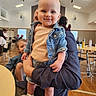 baby, child, denim_jacket, shorts, sandals, smile, indoor, person, adult, holding, people, room, table, chair, wood_floor, window, casual_clothing, event, gathering, happy