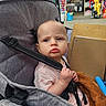 baby, stroller, child, face, pout, clothing, hand, safety_strap, seat, shopping_area, background, hats, toys, blur, person, portrait, expression, indoor, colorful, cute