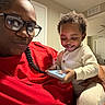 adult, baby, child, glasses, indoor, living_room, phone, smile, red_shirt, braid, curly_hair, white_clothing, holding, person, home, casual, joy, family, portrait, happy