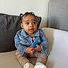 baby, child, denim_jacket, blue_toy, pigtails, couch, indoor, curious_expression, beige_pants, white_shoes, sitting, young_child, portrait, cute, infant, casual_clothing, home, cozy, happy, small_hands