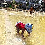 Anaya a rejoint le concours — aidez-le/la à gagner de superbes lots ! child, water, fountain, playing, outdoor, sunlight, splash, water_jet, urban, building, cap, red_shirt, shorts, summer, fun, happy, boy, reflection, pavement, daylight