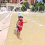 Anaya participe au concours pour gagner de l'argent avec cette photo : toddler, child, water, fountain, red_clothing, blue_hat, outdoor, summer, play, splash, urban, buildings, sunny, water_arc, standing, puddle, funny_face, daylight, young_child, happy