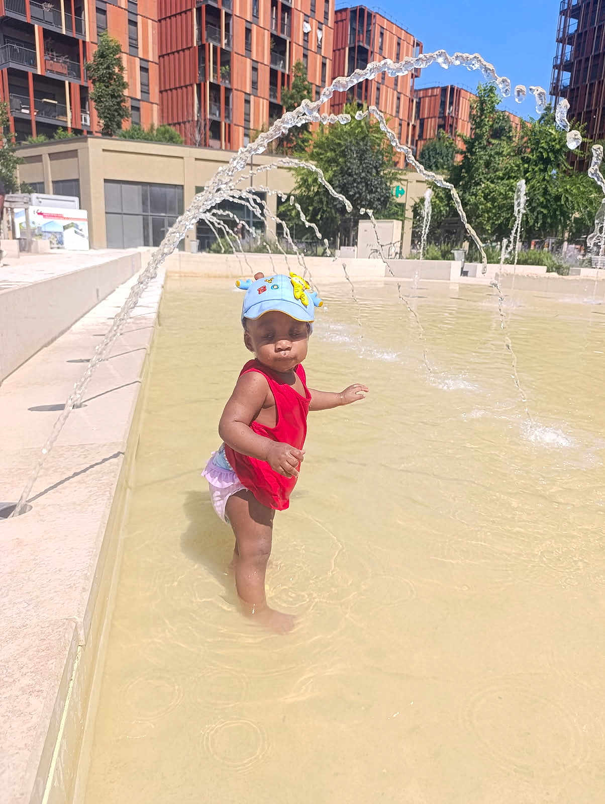 Anaya participe au concours pour gagner de l'argent avec cette photo : toddler, child, water, fountain, red_clothing, blue_hat, outdoor, summer, play, splash, urban, buildings, sunny, water_arc, standing, puddle, funny_face, daylight, young_child, happy