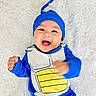 Andy is registered to the contest to win money with this photo: baby, smiling, blue_costume, yellow, happy, infant, lying_down, fluffy_background, cute, child, headwear, motion_blur, portrait, young_child, playful, costume, indoors, bright_colors, fun, expression