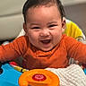baby, child, smiling, orange_clothing, face, indoor, toy, baby_walker, happy, cute, infant, playful, colorful, closeup, person, portrait, seat, fun, expression, young
