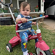 Asher joined the competition — help win amazing prizes! toddler, child, tricycle, radio_flyer, outdoor, grass, overalls, white_shoes, handlebars, porch, house, american_flag, plants, window, door, concrete, sidewalk, red, playful, curious