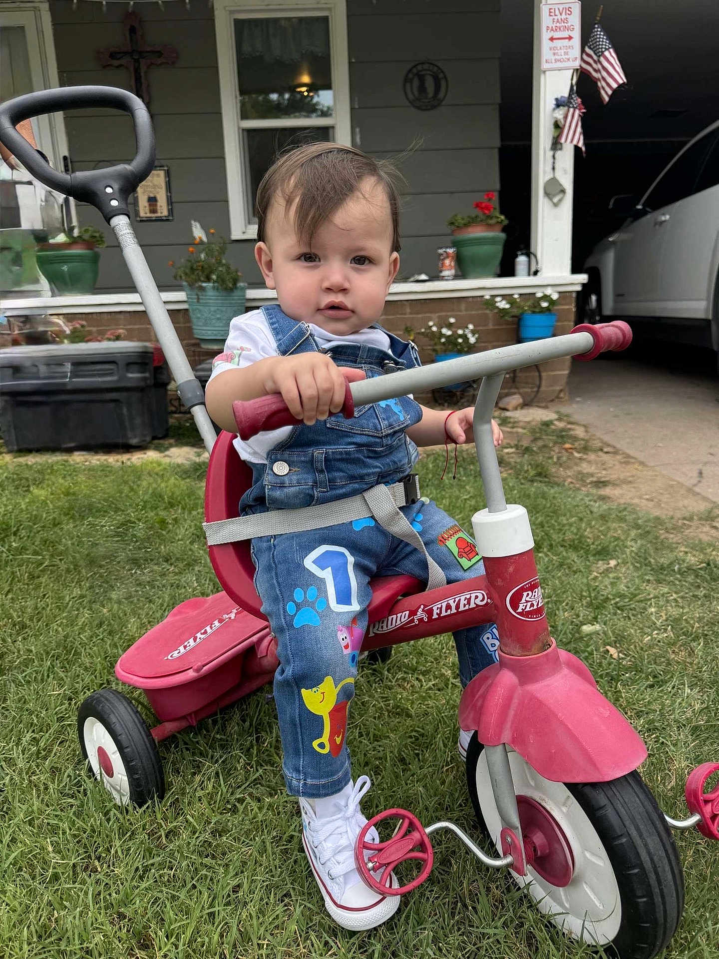 Asher joined the competition — help win amazing prizes! toddler, child, tricycle, radio_flyer, outdoor, grass, overalls, white_shoes, handlebars, porch, house, american_flag, plants, window, door, concrete, sidewalk, red, playful, curious