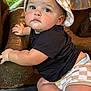 baby, child, face, drool, hat, black_shirt, diaper, checkered_pattern, bronze_sculpture, hand, leg, curious_expression, outdoor, greenery, skin, close_up, portrait, baby_clothing, infant, cute