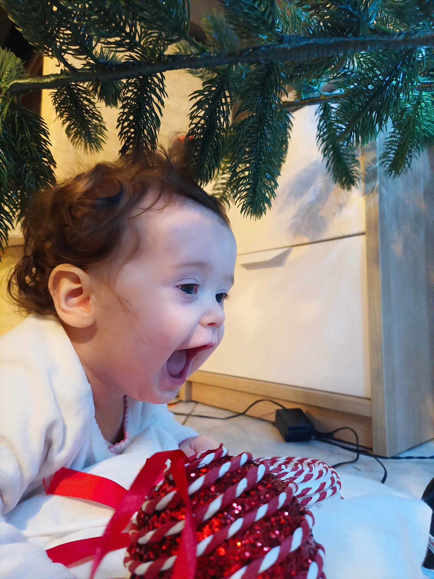 Yüna participe au concours pour gagner de l'argent avec cette photo : baby, child, curly_hair, christmas_tree, ornament, red_ribbon, indoor, floor, excitement, holiday, decorations, smile, happy, festive, closeup, toddler, face, mouth_open, candid, celebration
